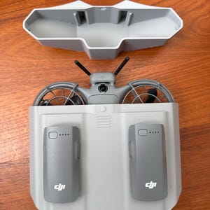 Puede incluir: Un conjunto de dron y estación de carga grises. El dron tiene dos hélices, dos antenas y una cámara. La estación de carga tiene dos ranuras para baterías y el logotipo de DJI. El conjunto está sobre una superficie de madera.