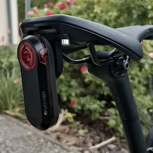 Puede incluir: Una luz trasera de bicicleta Garmin negra, sujeta a un sillín de bicicleta negro. La luz tiene una lente roja y el logotipo de Garmin es visible. El sillín está etiquetado como "BODY. GEOMETRY". El cuadro de la bicicleta es negro.