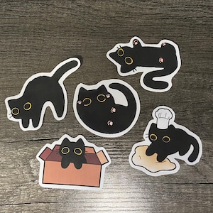 May include: Five black cat stickers with yellow eyes. The cats are in different poses, including sitting, lying down, and standing. One cat is wearing a chef's hat.