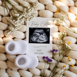 Könnte beinhalten: Eine Schwangerschaftsankündigung mit einem Ultraschallbild, weißen gehäkelten Babyschuhen und zarten weißen Blumen. Auf der Ankündigungskarte steht "Baby Chloe" und "DUE AUG 2026."