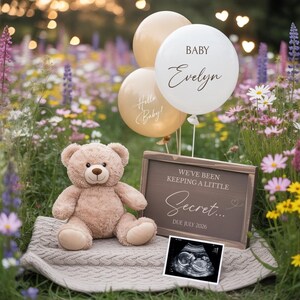 Könnte beinhalten: Ein Teddybär sitzt auf einer Decke neben einem Schild mit der Aufschrift "Wir haben ein kleines Geheimnis... Fälligkeit Juli 2026." Drei Ballons sagen "Hallo Baby!" und "Baby Evelyn." Ein Ultraschallbild ist ebenfalls vorhanden.