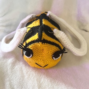 May include: A crocheted bee-shaped bag with black and yellow stripes, white crocheted handles, and black eyes.