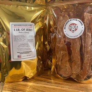 May include: Two gold-colored bags of chicken jerky for pets. One bag has a label with text including "BUTCHER BLOCK 4 PETS" and "1 LB. OF JERKY." The other bag has a circular sticker with a dog illustration and the text "100% CHICKEN JERKY."