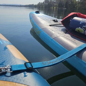 May include: A blue and grey inflatable stand-up paddleboard with the text Body Glove printed on the side. A blue nylon strap with the text SUPmate printed on it connects to the board. A dark blue water bottle with stickers sits on the deck.