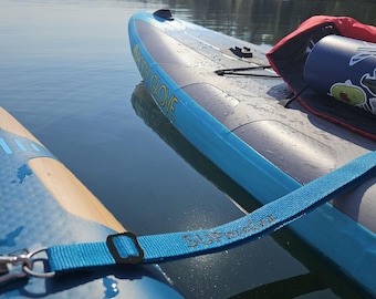 May include: A blue and grey inflatable stand-up paddleboard with the text Body Glove printed on the side. A blue nylon strap with the text SUPmate printed on it connects to the board. A dark blue water bottle with stickers sits on the deck.