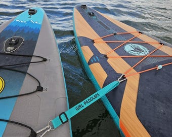 Puede incluir: Dos tablas de paddle surf flotando en el agua. Una tabla tiene un diseño azul y gris con una escena de montaña. La otra tiene un patrón de vetas de madera con cuerdas elásticas naranjas. Una correa turquesa con el texto "GIRL PADDLERS" conecta las tablas.