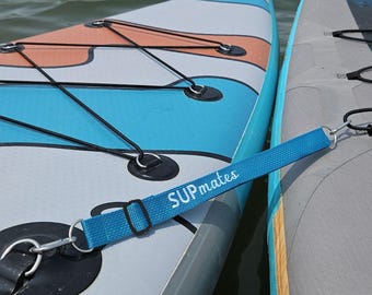 Könnte beinhalten: Zwei Stand-Up-Paddleboards, eines blau und weiß, das andere grau. Ein blaues Band mit dem Text "SUPmates" verbindet die Boards. Die Boards sind auf dem Wasser, mit schwarzen Bungee-Kordeln und Metallringen.