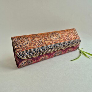 May include: A rectangular clutch bag featuring a woven fabric with a floral pattern in shades of orange, deep red, and gold. The flap is accented with a dark blue horizontal border, and the overall design displays intricate circular motifs.