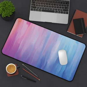 May include: A blue and pink abstract design mouse pad with a white computer mouse on top. The mouse pad is on a black surface with a laptop, a notebook, and a cup of coffee.