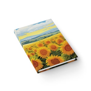 May include: A notebook with a colorful illustration of a field of sunflowers. The background shows a blue sky with white clouds and a distant landscape.