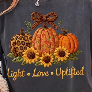Puede incluir: Camiseta gris oscuro con un diseño de tres calabazas y girasoles. Las calabazas tienen diferentes patrones, incluyendo estampado de leopardo y floral. El texto "Light • Love • Uplifted" está impreso en dorado debajo.