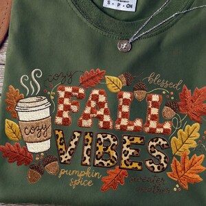 Puede incluir: Camiseta verde oliva con diseño bordado "Fall Vibes". El diseño incluye una taza de café, hojas de otoño y las palabras "cozy", "blessed", "pumpkin spice" y "sweater weather". También se ven un collar plateado y un sombrero marrón.