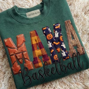 Puede incluir: Sudadera verde con la palabra "MAMA" en una mezcla de texturas: baloncesto, cuadros y tela con temática de Halloween. La palabra "Basketball" está bordada en cursiva. La marca "Cotton & Co" está en el cuello.