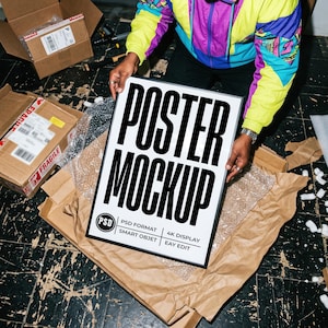Puede incluir: Una maqueta de póster enmarcada en negro con las palabras "POSTER MOCKUP" en letras blancas en negrita. El póster está sostenido por una persona. El póster está sobre papel de embalaje marrón y plástico de burbujas. Hay cajas de cartón en el fondo.
