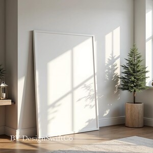 Puede incluir: Un marco de fotos blanco con un lienzo en blanco, de pie sobre un suelo de madera en una habitación con un pequeño pino en una maceta marrón. La habitación tiene paredes blancas y una ventana con la luz del sol entrando.