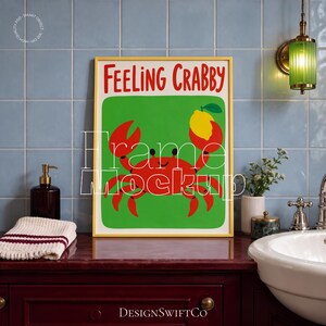 Puede incluir: Un cuadro rectangular con marco amarillo que muestra un cangrejo rojo sobre un fondo verde sosteniendo un limón amarillo, con el texto rojo "Feeling Crabby" en la parte superior. La obra se exhibe en un baño con azulejos azules.