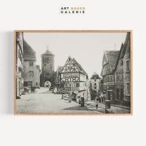 Op de afbeelding: Zwart-wit ingelijste prent van een Europees stadsplein. De afbeelding toont gebouwen met puntdaken, een klokkentoren en geplaveide straten. Het kunstwerk zit in een houten lijst en heeft bovenaan de tekst "ART BAUER GALERIE".