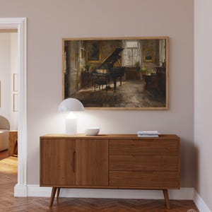 May include: A wooden sideboard with two doors and three drawers. The sideboard is a light brown color and has a white lamp and a bowl on top. A framed painting of a piano in a room is hanging on the wall above the sideboard.