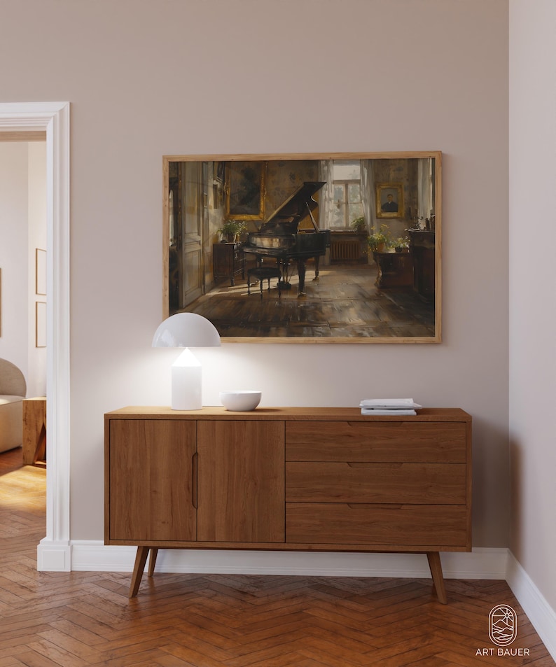 May include: A wooden sideboard with two doors and three drawers. The sideboard is a light brown color and has a white lamp and a bowl on top. A framed painting of a piano in a room is hanging on the wall above the sideboard.