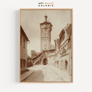 Op de afbeelding: Een zwart-witfoto van een smalle straat in een Europese stad. De straat is omzoomd met oude gebouwen, en in de verte is een hoge toren te zien. De foto is ingelijst in een houten lijst.