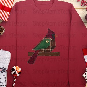 May include: A burgundy sweatshirt with an appliqued cardinal bird design. The bird is made of green, plaid, and burgundy fabric with a button detail. The sweatshirt is surrounded by festive decorations, including a gift box and baubles.