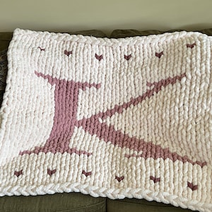 May include: A white knitted blanket with a large pink letter K in the center. The blanket has a braided border and small pink hearts scattered throughout.