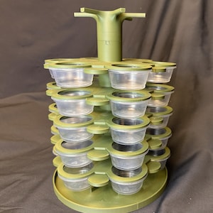 May include: A green plastic spice rack with multiple tiers of small, clear containers. The rack has a central support structure and a circular base. The containers are arranged in a circular pattern around the central support.