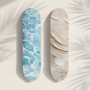 May include: Two skateboards with artistic designs. One features a blue water pattern, the other a textured beige design. Both have four black screw holes. The boards are displayed against a white wall with palm leaf shadows.