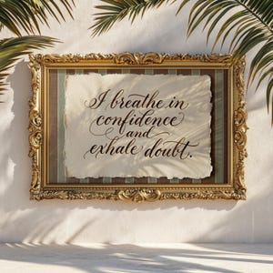 May include: A framed artwork with the text "I breathe in confidence and exhale doubt." The ornate gold frame surrounds a piece of paper with a textured edge, set against a striped background. Palm fronds are visible in the background.