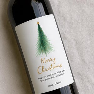 May include: A dark wine bottle with a white label featuring a watercolor Christmas tree design. The label includes the text "Merry Christmas" in gold script, along with a holiday message and a name.