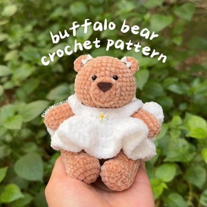 Op de afbeelding: Een gehaakte bruine teddybeer die een witte jurk draagt met een klein wit bloemetje. De beer heeft een bruine neus en zwarte ogen. De tekst "buffalo bear crochet pattern" staat boven de beer. De tekst "@crochetingbirds" staat onder de beer.