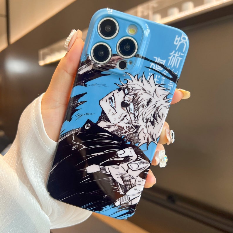 K&ouml;nnte beinhalten: Eine blaue Handyh&uuml;lle mit einer Schwarzwei&szlig;illustration eines Charakters aus dem Anime "Jujutsu Kaisen". Der Charakter hat stacheliges wei&szlig;es Haar und tr&auml;gt eine schwarze Jacke. Die H&uuml;lle hat einen Ausschnitt f&uuml;r das Kameraobjektiv und den japanischen Text "呪術廻戦", der sich mit "Jujutsu Kaisen" &uuml;bersetzen l&auml;sst.