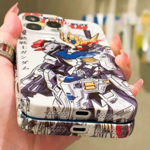 Puede incluir: Una funda de tel&eacute;fono blanca con una ilustraci&oacute;n de estilo anime de un robot. La funda tiene un relieve de c&aacute;mara elevado y un texto negro que dice "機動戦士ガンダム".