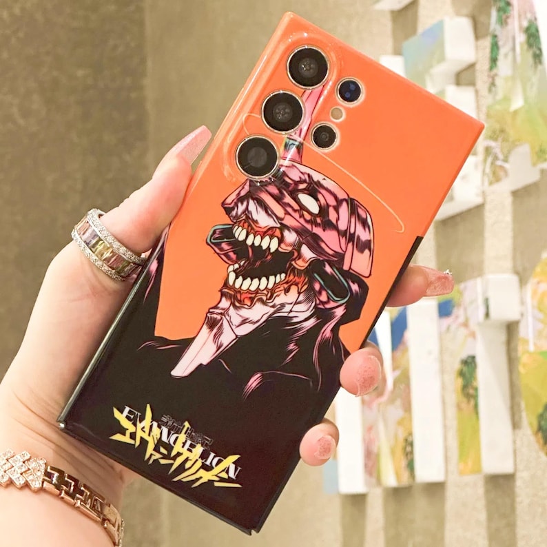 Może przedstawiać: Pomarańczowe etui na telefon z czarno-r&oacute;żowym wzorem postaci z anime. Postać ma duże usta z ostrymi zębami i nosi hełm. Etui ma wystający aparat z pięcioma obiektywami. Na etui nadrukowany jest tekst "EVANGELION" w japońskich znakach.