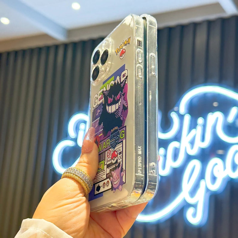 Może przedstawiać: Przezroczyste etui na telefon z kolorową grafiką postaci Pokemon, Genga, z Ligi Pokemon. Etui jest przeznaczone do iPhone'a 13 Pro Max.
