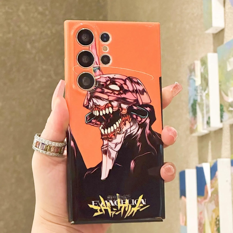 Może przedstawiać: Pomarańczowe etui na telefon z czarno-r&oacute;żowym wzorem postaci z anime. Postać ma duże usta z ostrymi zębami i pochodzi z anime Evangelion. Etui ma wystający element z trzema obiektywami aparatu.