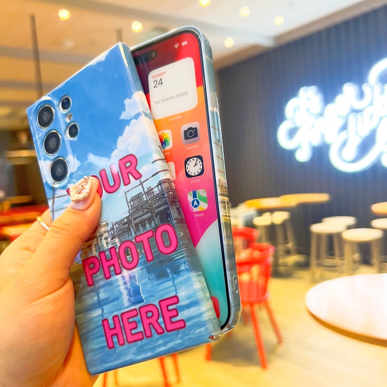 May include: A blue phone case with a white and blue sky background. The case has the text "YOUR PHOTO HERE" in pink letters. The phone case is being held in a hand.