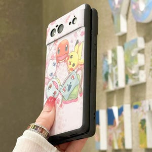 May include: A pink and white phone case with a black border. The case features a design of cartoon characters from the Pokemon franchise, including Bulbasaur, Charmander, Squirtle, and Pikachu. The characters are surrounded by pink cherry blossoms.
