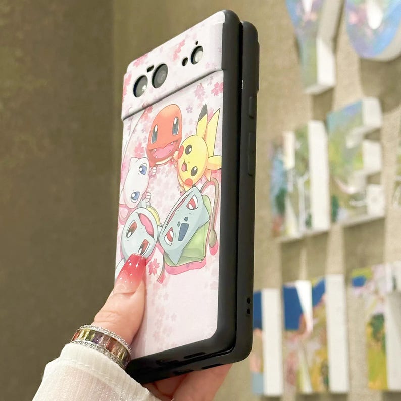 May include: A pink and white phone case with a black border. The case features a design of cartoon characters from the Pokemon franchise, including Bulbasaur, Charmander, Squirtle, and Pikachu. The characters are surrounded by pink cherry blossoms.