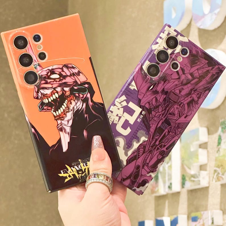 Może przedstawiać: Dwa etui na telefon z wzorami inspirowanymi anime. Jedno etui przedstawia fioletowego i czarnego robota z japońskim tekstem. Drugie etui przedstawia czerwonego i czarnego potwora na jasno pomarańczowym tle. Oba etui mają wycięcie na aparat wieloobiektywowy.