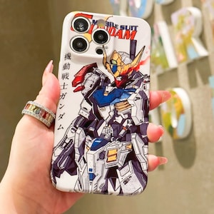 Puede incluir: Una funda blanca para iPhone con una ilustraci&oacute;n colorida de un robot Gundam. La funda tiene un recorte para el objetivo de la c&aacute;mara y el texto "Mobile Suit Gundam" est&aacute; impreso en la parte superior de la funda.