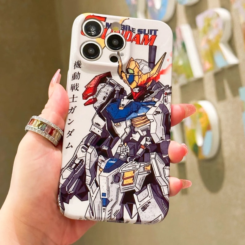 Puede incluir: Una funda blanca para iPhone con una ilustraci&oacute;n colorida de un robot Gundam. La funda tiene un recorte para el objetivo de la c&aacute;mara y el texto "Mobile Suit Gundam" est&aacute; impreso en la parte superior de la funda.