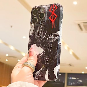 Może przedstawiać: Białe etui na iPhone z czarno-białą ilustracją postaci z anime Berserk. Postać jest ubrana w zbroję i trzyma miecz. Etui zawiera r&oacute;wnież czerwony symbol "Znaku Ofiary".
