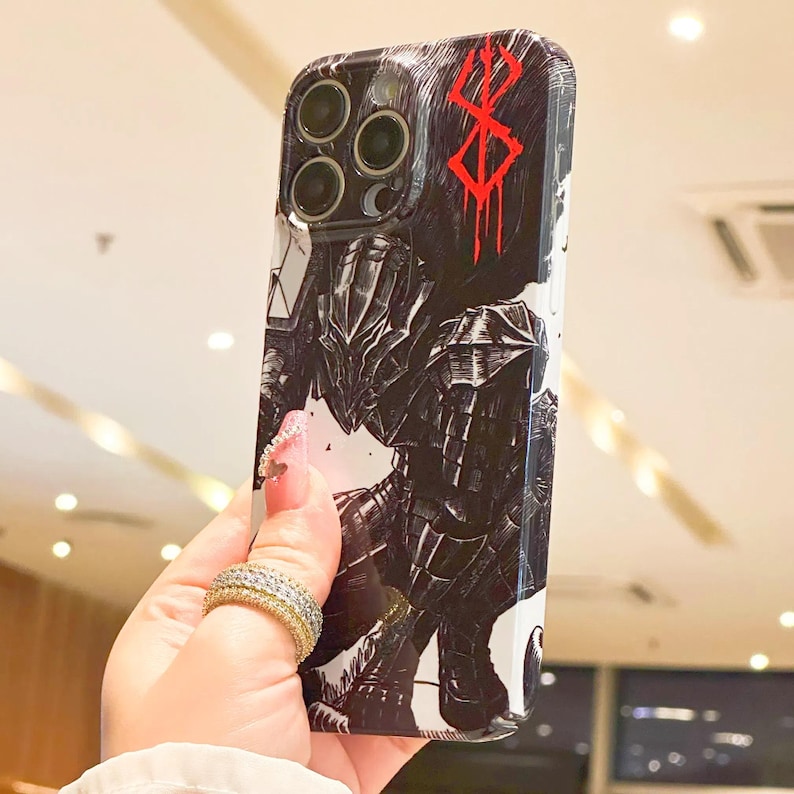Może przedstawiać: Białe etui na iPhone z czarno-białą ilustracją postaci z anime Berserk. Postać jest ubrana w zbroję i trzyma miecz. Etui zawiera r&oacute;wnież czerwony symbol "Znaku Ofiary".