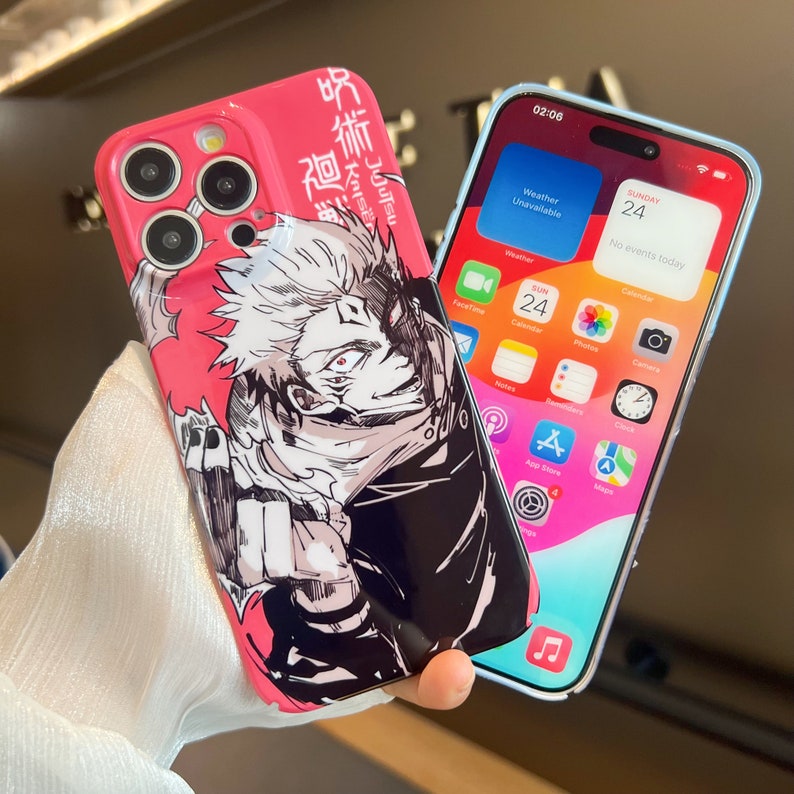 K&ouml;nnte beinhalten: Eine rosa iPhone-H&uuml;lle mit einer Schwarzwei&szlig;-Illustration eines Charakters aus dem Anime Jujutsu Kaisen. Die H&uuml;lle hat japanischen Text auf der R&uuml;ckseite. Die Handyh&uuml;lle wird neben einer durchsichtigen iPhone-H&uuml;lle mit blauem Rand gezeigt.