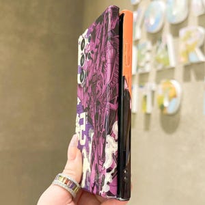 Może przedstawiać: Czarno-fioletowe etui na telefon z komiksowym wzorem. Etui ma czarny i pomarańczowy boczny panel.