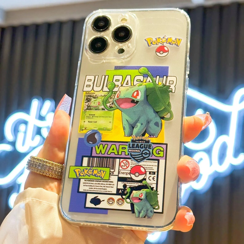 Może przedstawiać: Przezroczyste etui na telefon z wzorem Pokemon Bulbasaur. Etui przedstawia zielonego i niebieskiego Bulbasaura z napisem "Bulbasaur" i "Pokemon" na etui. Etui zawiera r&oacute;wnież naklejkę z logo Pokemon League z napisem "Taiwan League Regional".