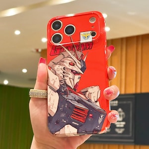May include: A bright red smartphone case featuring a detailed graphic of a white and blue Gundam robot. The word GUNDAM is printed in bold red letters above the robot illustration. The case includes cutouts for the camera lenses and a small emblem.