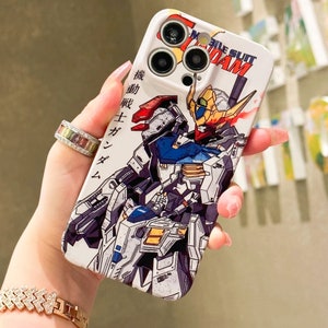 Puede incluir: Una funda blanca para iPhone con una ilustraci&oacute;n colorida de dos robots Gundam. La funda tiene un recorte para el objetivo de la c&aacute;mara y el texto "Mobile Suit Gundam" impreso en ella.