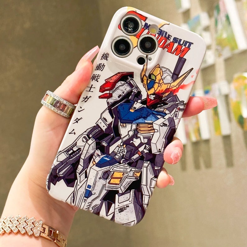 Puede incluir: Una funda blanca para iPhone con una ilustraci&oacute;n colorida de dos robots Gundam. La funda tiene un recorte para el objetivo de la c&aacute;mara y el texto "Mobile Suit Gundam" impreso en ella.
